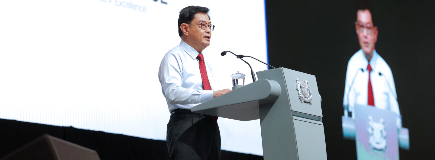 Man in shirt/tie at podium with Singapore crest, speaking to audience. Large screen behind shows his reflection.
